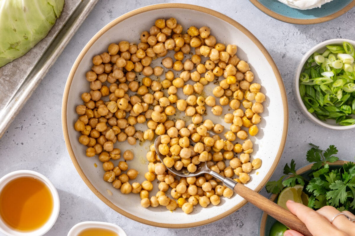 A bowl of seasoned chickpeas is being stirred with a spoon. Surrounding the bowl are small dishes of sauces, sliced green onions, fresh herbs, and part of a cabbage.