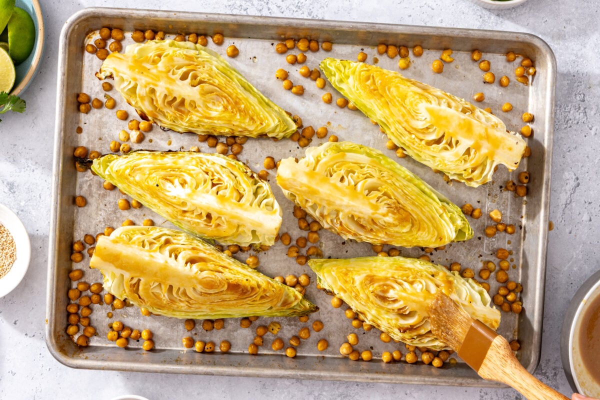 Six cabbage wedges and roasted chickpeas on a baking sheet. A hand brushes glaze on one cabbage wedge. Small bowls with lime, seeds, and herbs are nearby on a light countertop.
