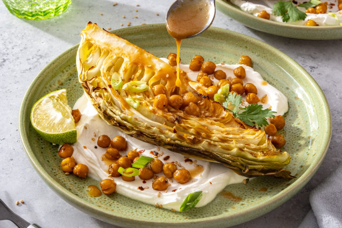 A roasted cabbage wedge on yogurt sauce, topped with crispy chickpeas, green onions, cilantro, and sauce being drizzled from a spoon. A lime wedge sits on the side of the green plate.