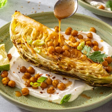 A roasted cabbage wedge on yogurt sauce, topped with crispy chickpeas, green onions, cilantro, and sauce being drizzled from a spoon. A lime wedge sits on the side of the green plate.