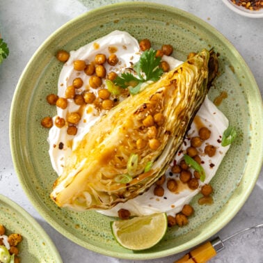 A charred wedge of cabbage on a plate with creamy sauce, roasted chickpeas, fresh cilantro, green onions, red pepper flakes, and a lime wedge. Other plates and ingredients surround the main dish.